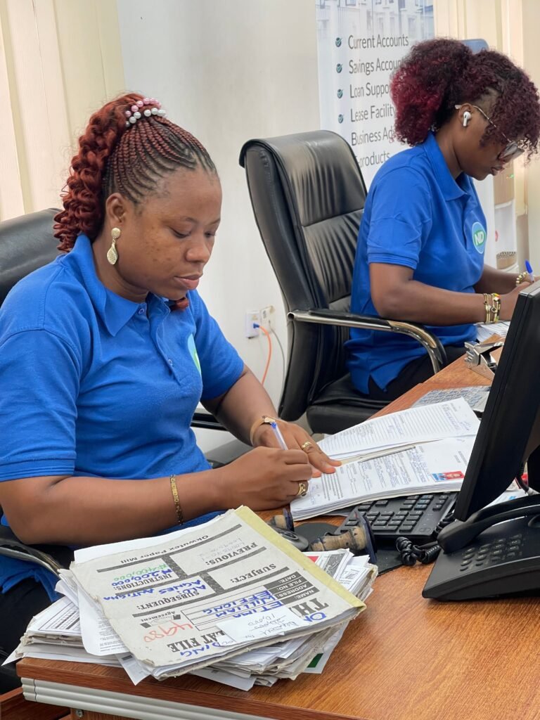 Two women in casual office attire working at desks in a business setting, reviewing documents.